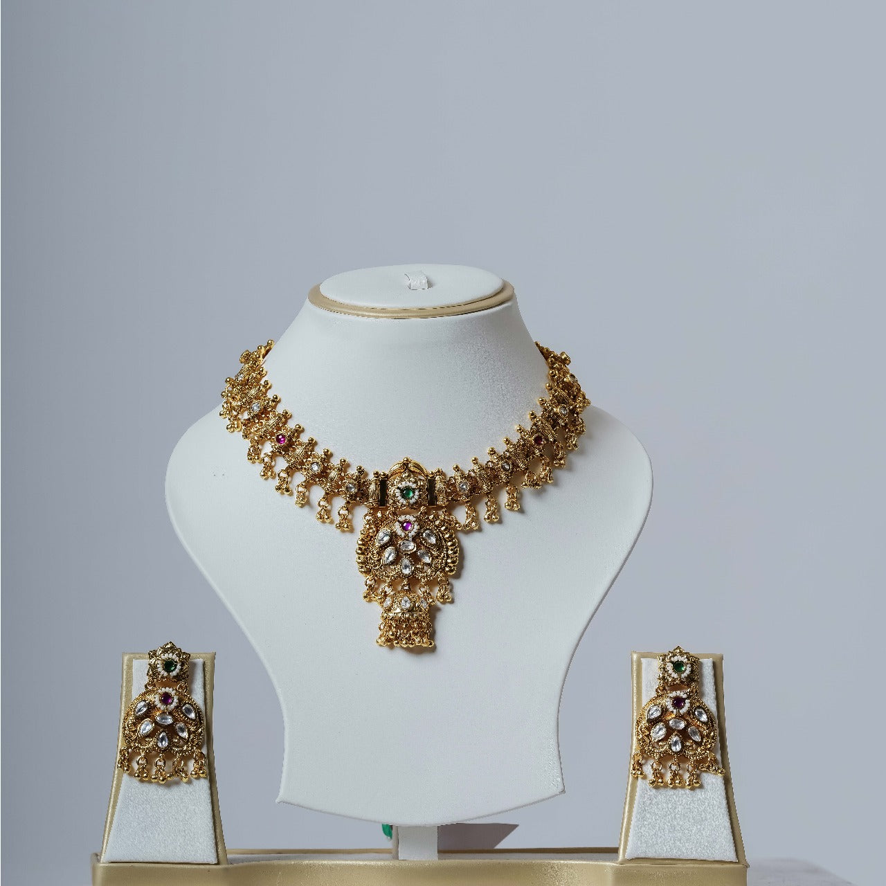 A lavish gold-toned Kundan necklace set with a detailed central pendant and multi-colored stones, accompanied by matching long drop earrings, displayed on white jewelry busts against a plain white background.