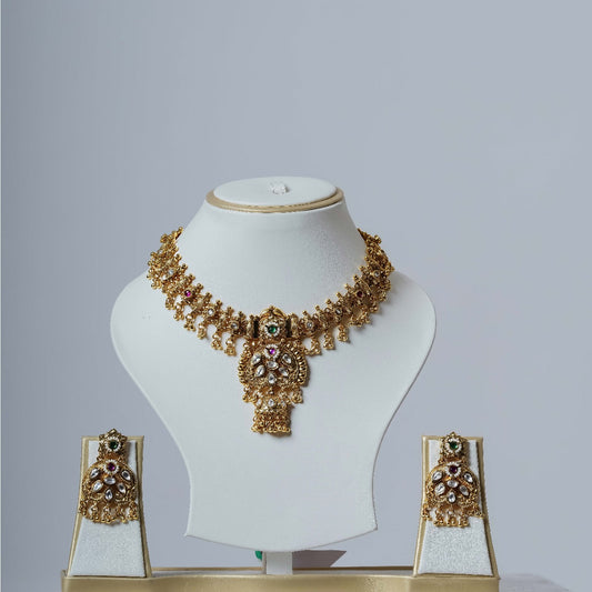 A lavish gold-toned Kundan necklace set with a detailed central pendant and multi-colored stones, accompanied by matching long drop earrings, displayed on white jewelry busts against a plain white background.