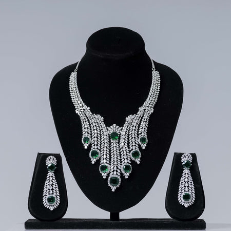 A lavish emerald and diamond statement bridal necklace set with a cascading design and prominent green gemstones, paired with matching long dangle earrings, displayed on black velvet jewelry busts against a neutral gray background.