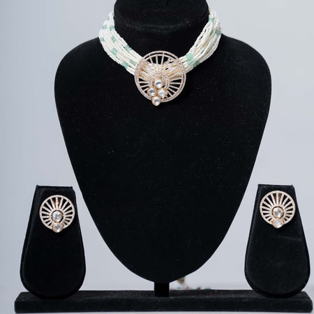A contemporary beaded choker necklace in white and light green with a central gold-toned medallion featuring sparkling stones, paired with matching stud earrings, displayed on black jewelry busts against a light gray background.