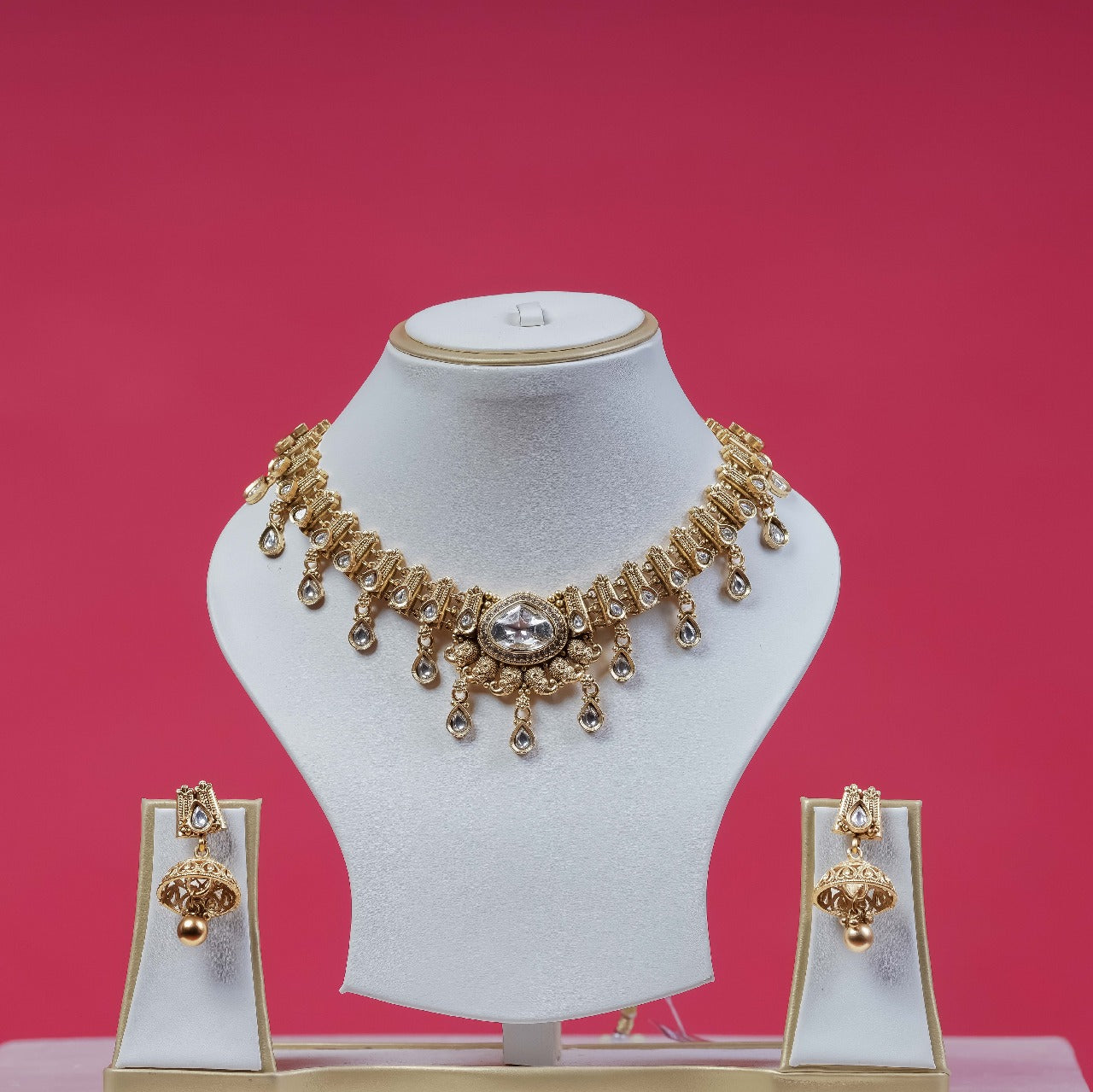 A traditional gold-toned polki necklace set with clear stones and dangling accents, paired with matching jhumka earrings, displayed on white jewelry busts against a vibrant pink background.