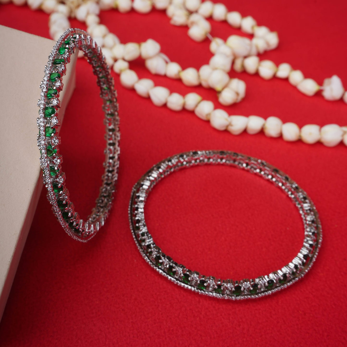 Silver bangles with emerald green and white stones on red fabric background - Revaaj