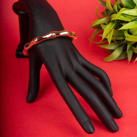 Gold-tone hinged bangle bracelet with a bamboo-like textured surface, next to a white block and a white beaded necklace on a red background - Revaaj