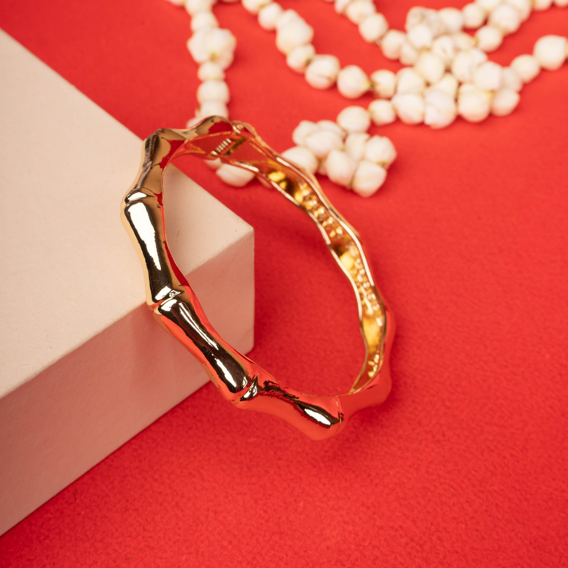 Gold-tone hinged bangle bracelet with a bamboo-like textured surface, next to a white block and a white beaded necklace on a ground background - Revaaj