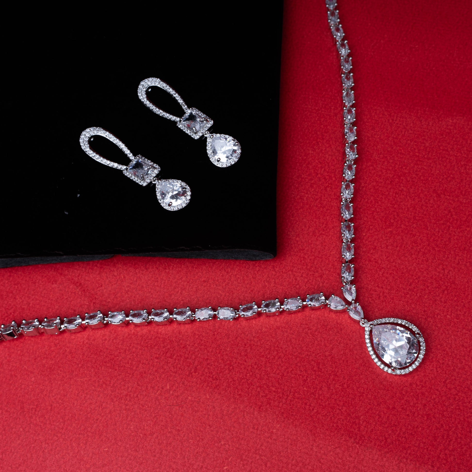 Silver Coloured necklace and earring set with teardrop and square diamond accents on red background