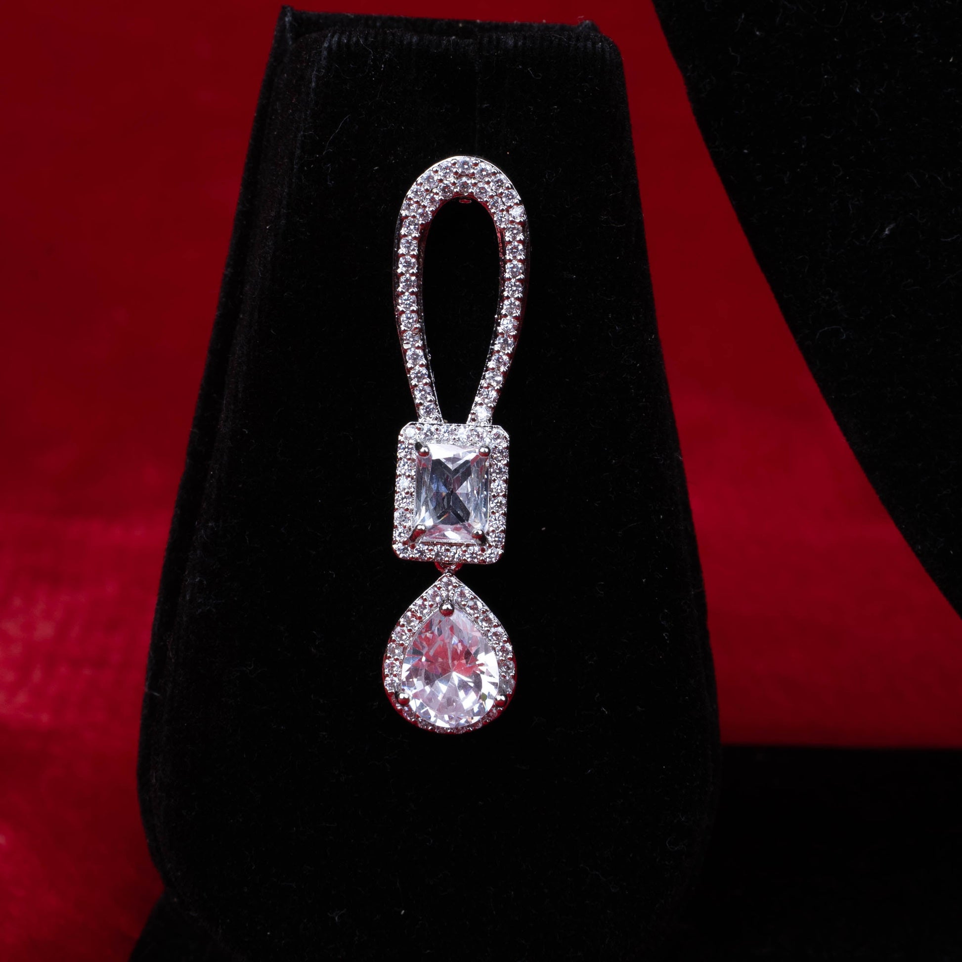 Earring with teardrop-shaped clear stone and rectangular clear stone in metallic setting on black velvet with red background