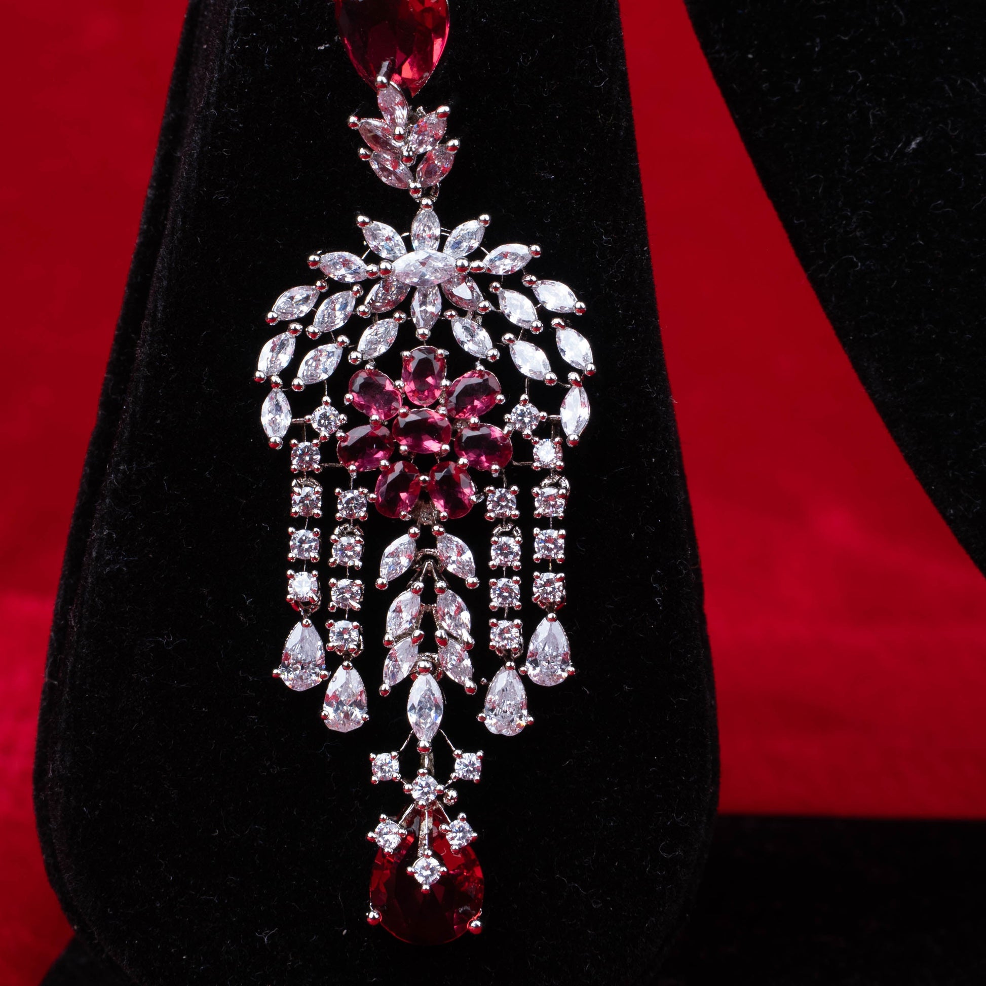 Earring with red teardrop stones, floral centerpiece, and cascading clear stone accents on black velvet with red background