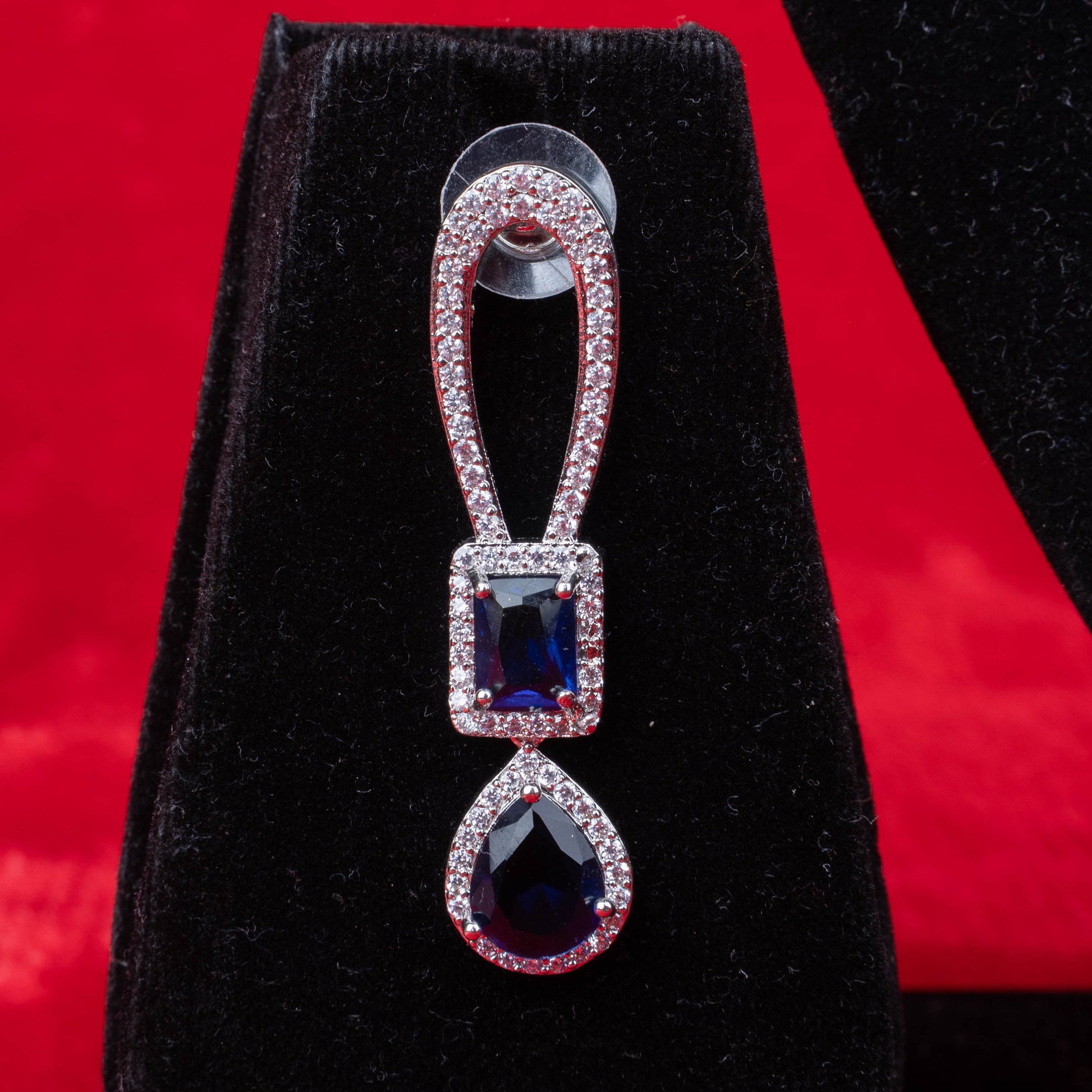 Earring with blue rectangular and teardrop stones in halo setting on black velvet with red background