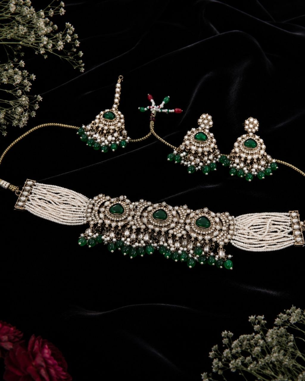 Emerald green kundan choker necklace set with white pearl tassels, matching drop earrings and maang tikka displayed on beige silk fabric with flowers 3