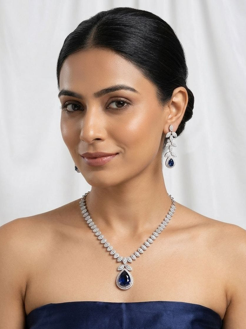 Silver necklace with large blue teardrop pendant surrounded by clear stones and matching leaf drop earrings on white fabric 2