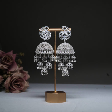 Sterling silver plated jhumka earrings with intricate filigree design and cascading bell charms, adorned with sparkling cubic zirconia stones, displayed on a golden stand with a floral background