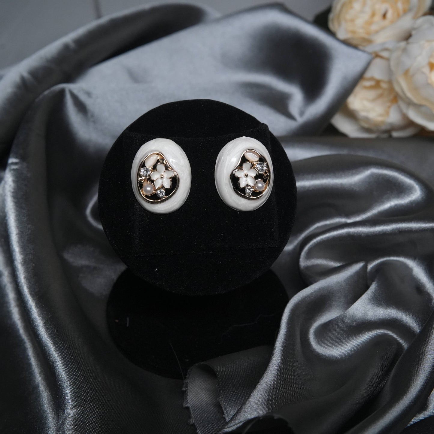 White enamel floral stud earrings with pearl and cubic zirconia accents, oval design displayed on a black velvet stand with silk background