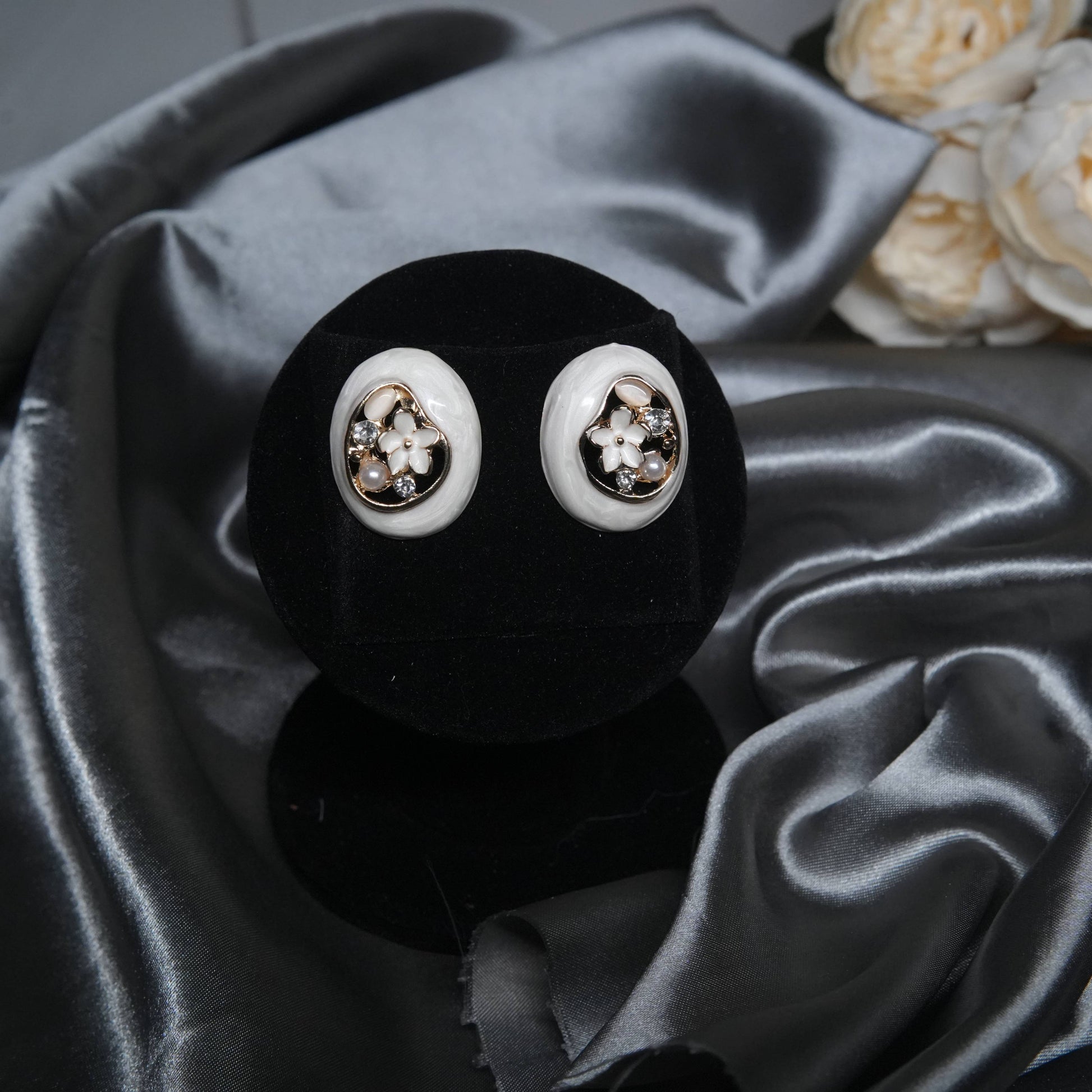 White enamel floral stud earrings with pearl and cubic zirconia accents, oval design displayed on a black velvet stand with silk background