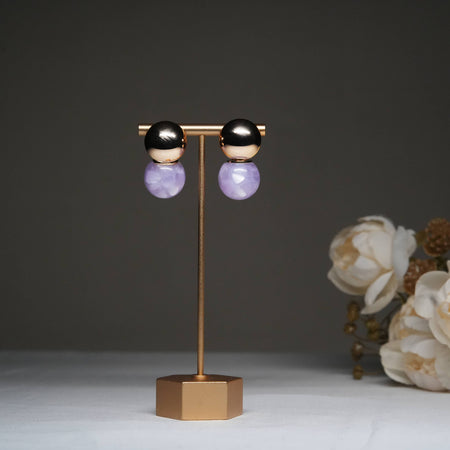 Dual tone metallic stud earrings with lavender stone drops displayed on a golden stand with floral backdrop.