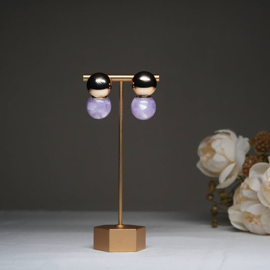 Dual tone metallic stud earrings with lavender stone drops displayed on a golden stand with floral backdrop.