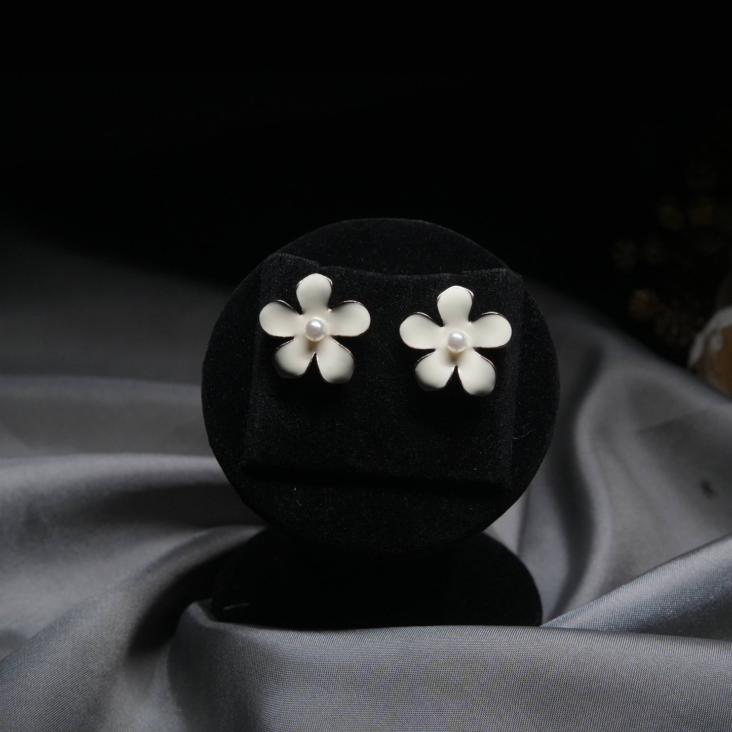White enamel floral stud earrings with pearl center displayed on black velvet stand.
