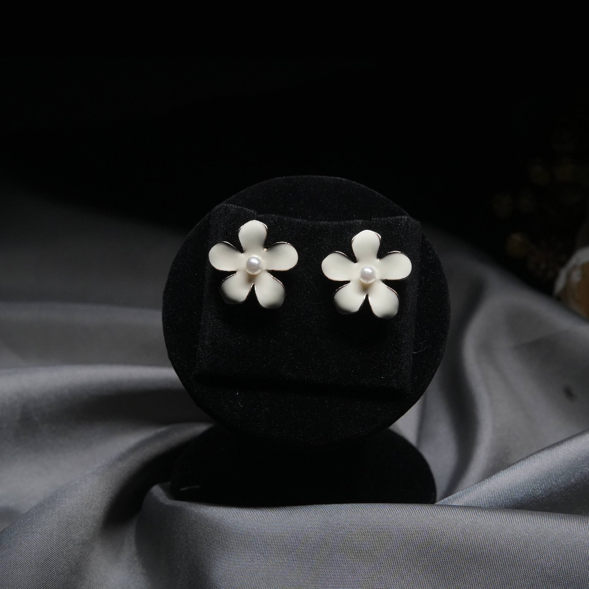 White enamel floral stud earrings with pearl center displayed on black velvet stand.
