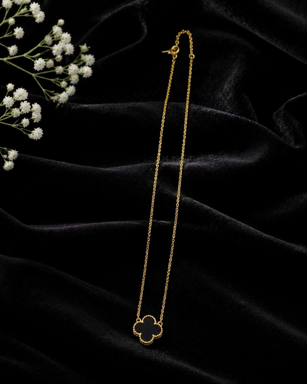 Delicate gold chain necklace with black enamel four-leaf clover pendant displayed on beige surface with white and pink flowers 3