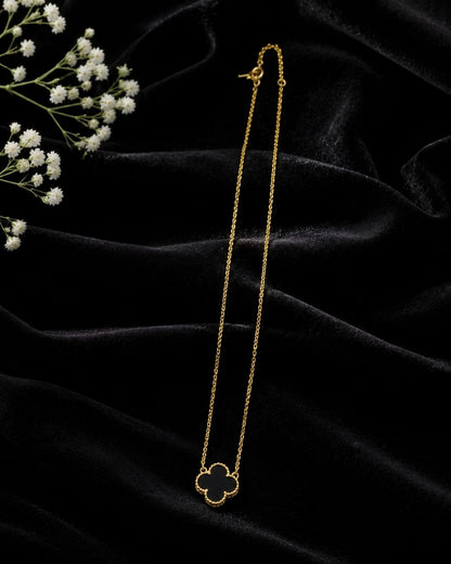 Delicate gold chain necklace with black enamel four-leaf clover pendant displayed on beige surface with white and pink flowers 3