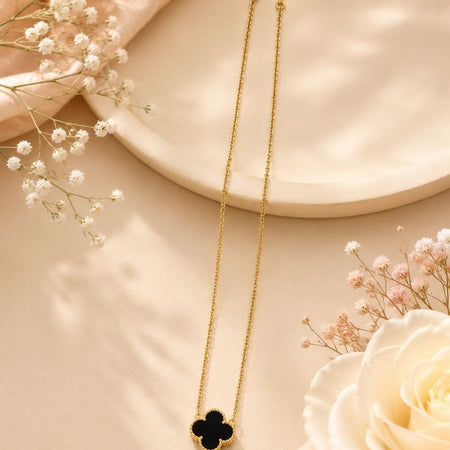 Delicate gold chain necklace with black enamel four-leaf clover pendant displayed on beige surface with white and pink flowers 1