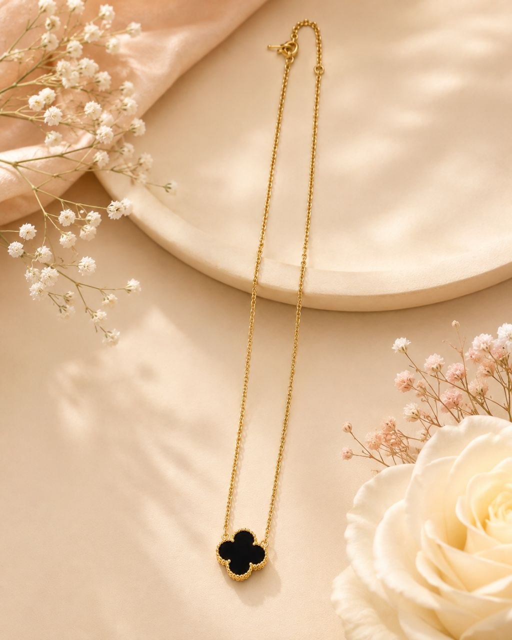 Delicate gold chain necklace with black enamel four-leaf clover pendant displayed on beige surface with white and pink flowers 1