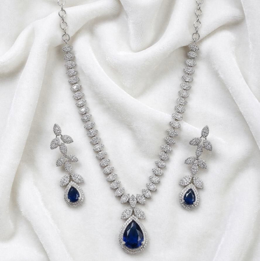 Silver necklace with large blue teardrop pendant surrounded by clear stones and matching leaf drop earrings on white fabric 1