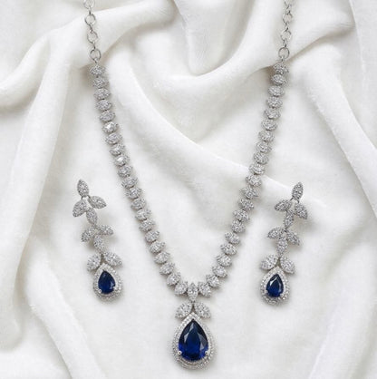 Silver necklace with large blue teardrop pendant surrounded by clear stones and matching leaf drop earrings on white fabric 1