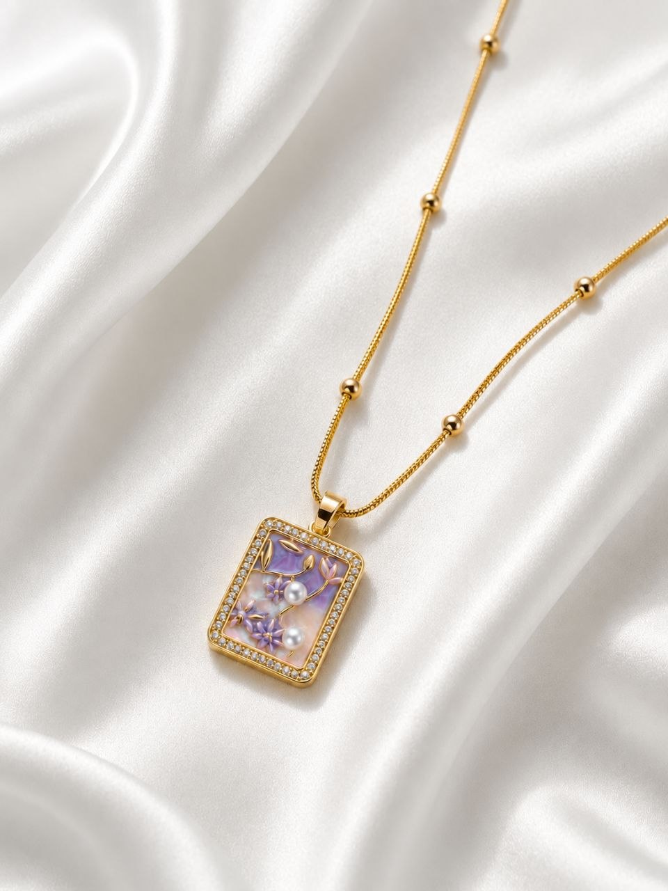 Gold beaded chain necklace with rectangular pendant featuring purple floral design, white pearls and crystal border on white satin fabric 1