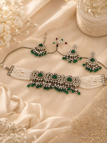 Emerald green kundan choker necklace set with white pearl tassels, matching drop earrings and maang tikka displayed on beige silk fabric with flowers 1