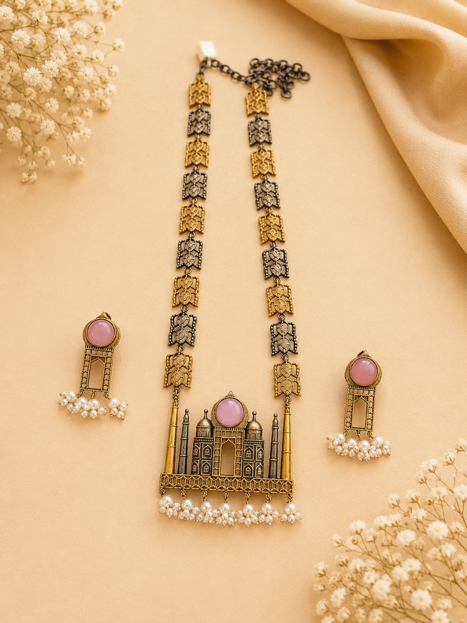 Antique gold and black Taj Mahal motif necklace with pink stone pendant and pearl drops alongside matching earrings on beige fabric with white flowers 1