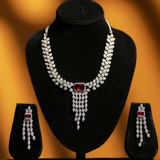Close-up of a luxurious diamond and ruby bridal necklace set with a leaf design and matching dangle earrings displayed on black velvet busts against a warm orange background.