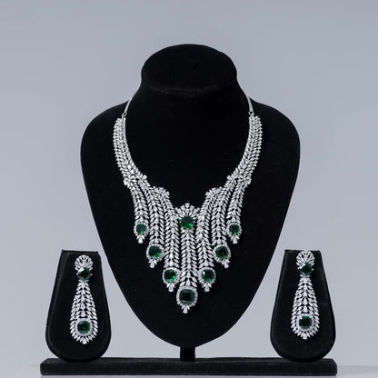 A lavish emerald and diamond statement bridal necklace set with a cascading design and prominent green gemstones, paired with matching long dangle earrings, displayed on black velvet jewelry busts against a neutral gray background.