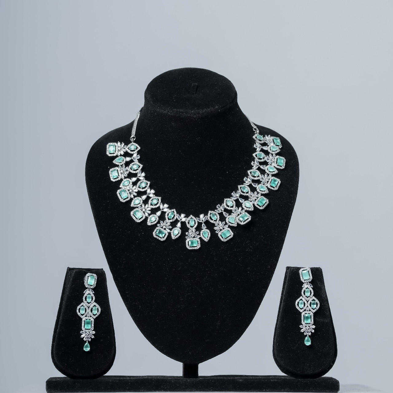 A dazzling emerald and diamond statement necklace set with matching drop earrings displayed on black velvet jewelry busts against a neutral gray background.