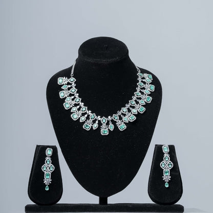 A dazzling emerald and diamond statement necklace set with matching drop earrings displayed on black velvet jewelry busts against a neutral gray background.