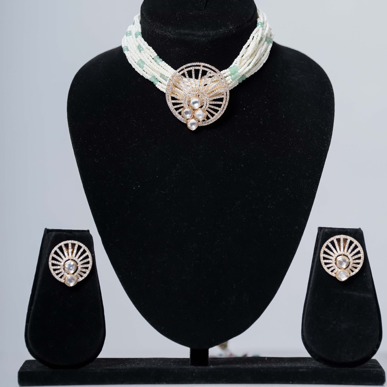 A contemporary beaded choker necklace in white and light green with a central gold-toned medallion featuring sparkling stones, paired with matching stud earrings, displayed on black jewelry busts against a light gray background.