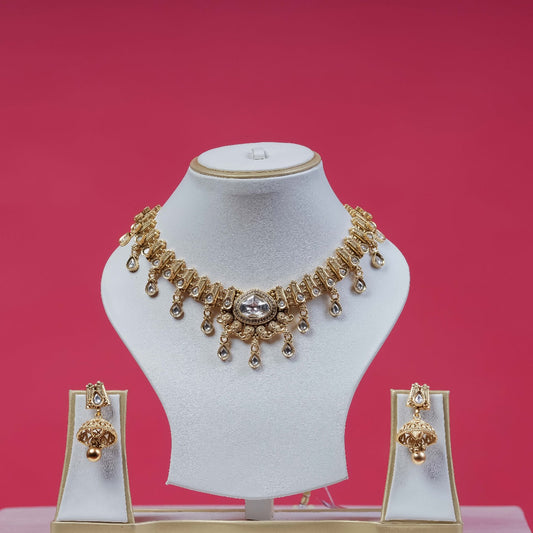 A traditional gold-toned polki necklace set with clear stones and dangling accents, paired with matching jhumka earrings, displayed on white jewelry busts against a vibrant pink background.