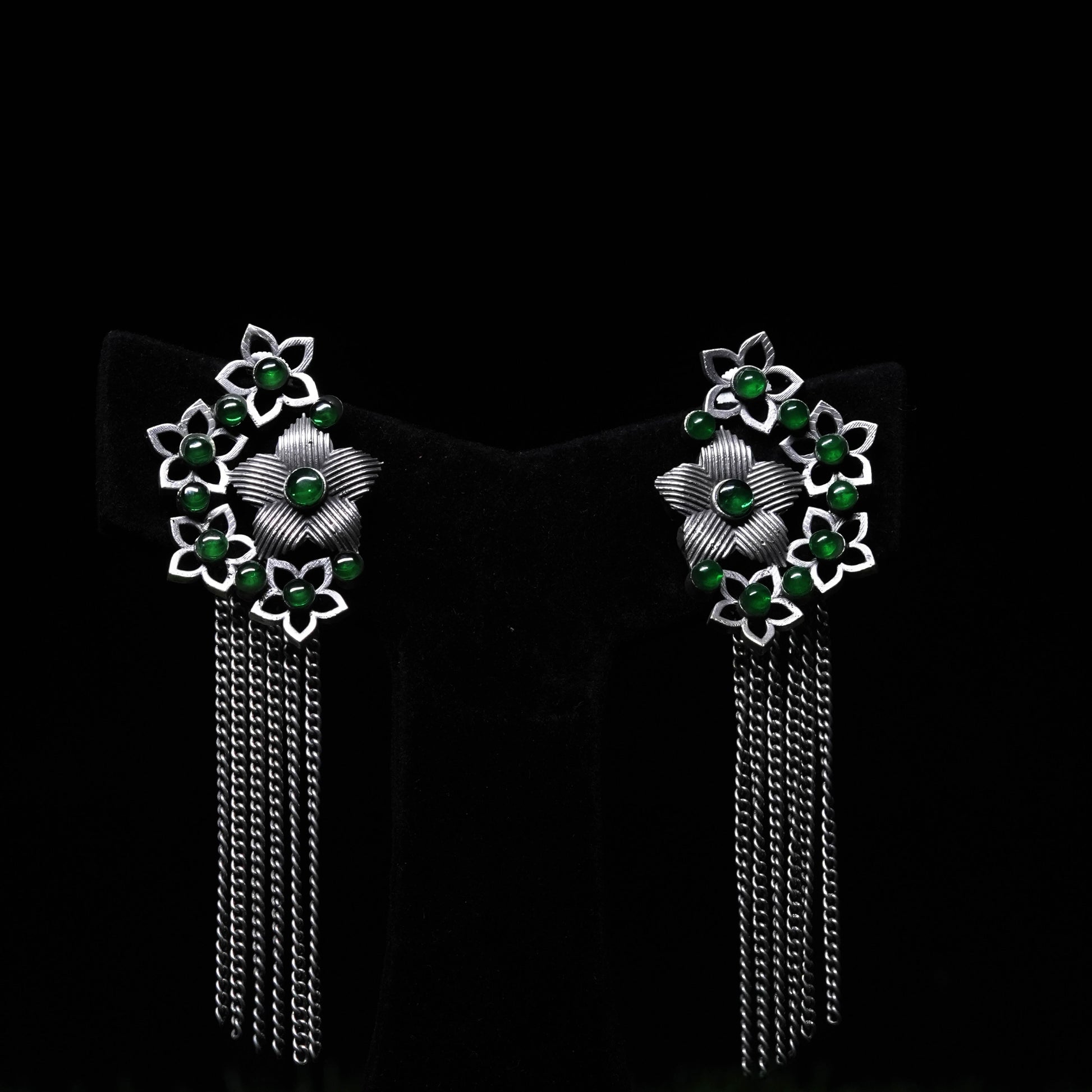 Oxidized floral tassel earrings with green bead accents and cascading chains displayed on black stand 