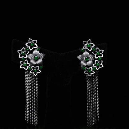 Oxidized floral tassel earrings with green bead accents and cascading chains displayed on black stand 