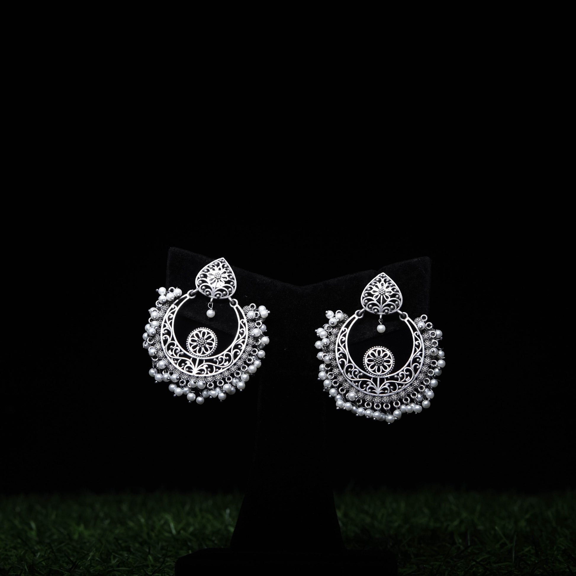 Oxidized silver chandbali earrings with intricate filigree and pearl bead accents displayed on black stand