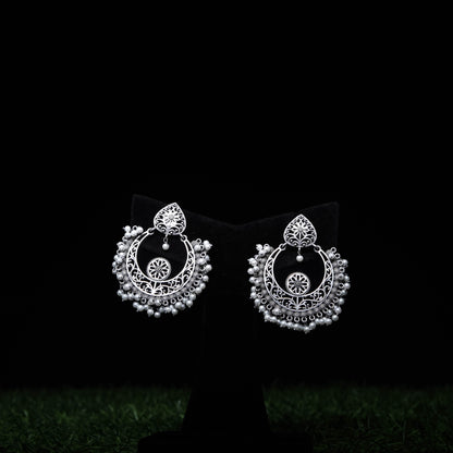Oxidized silver chandbali earrings with intricate filigree and pearl bead accents displayed on black stand