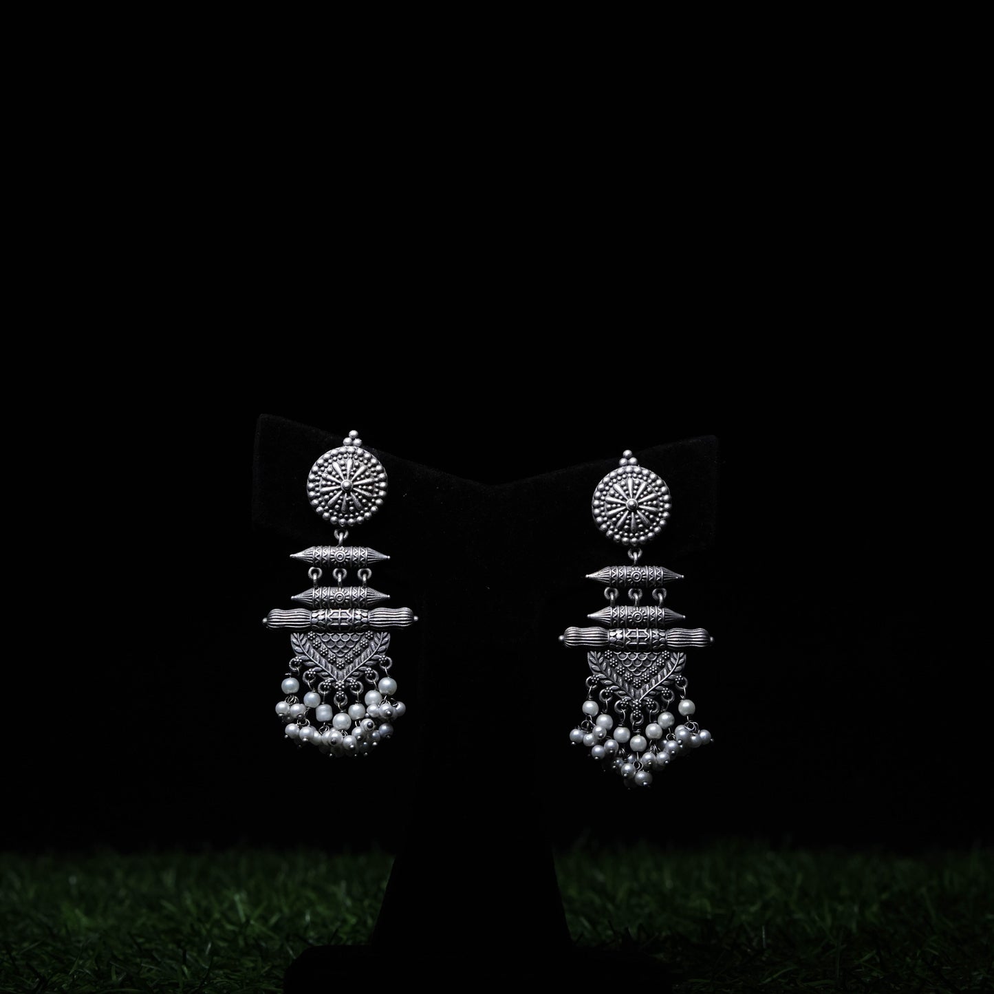 Oxidized silver tribal dangler earrings with intricate detailing and pearl drops displayed on black stand