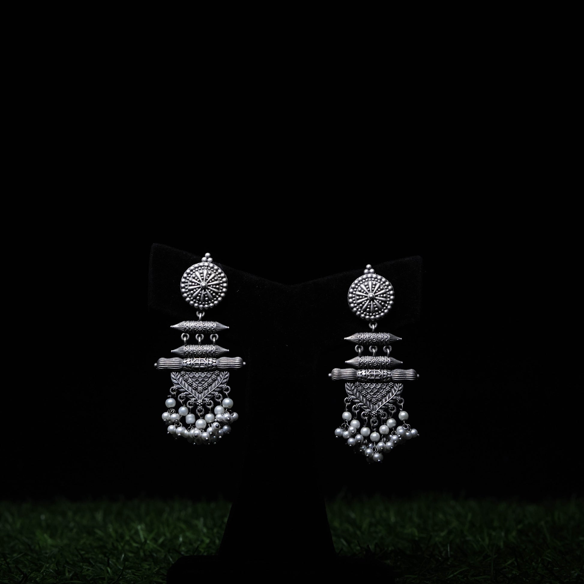 Oxidized silver tribal dangler earrings with intricate detailing and pearl drops displayed on black stand