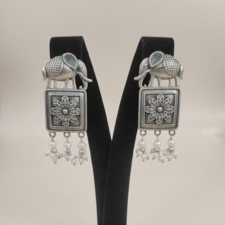 Oxidized silver earrings with elephant motifs, floral square design, and pearl drop detailing displayed on a black stand