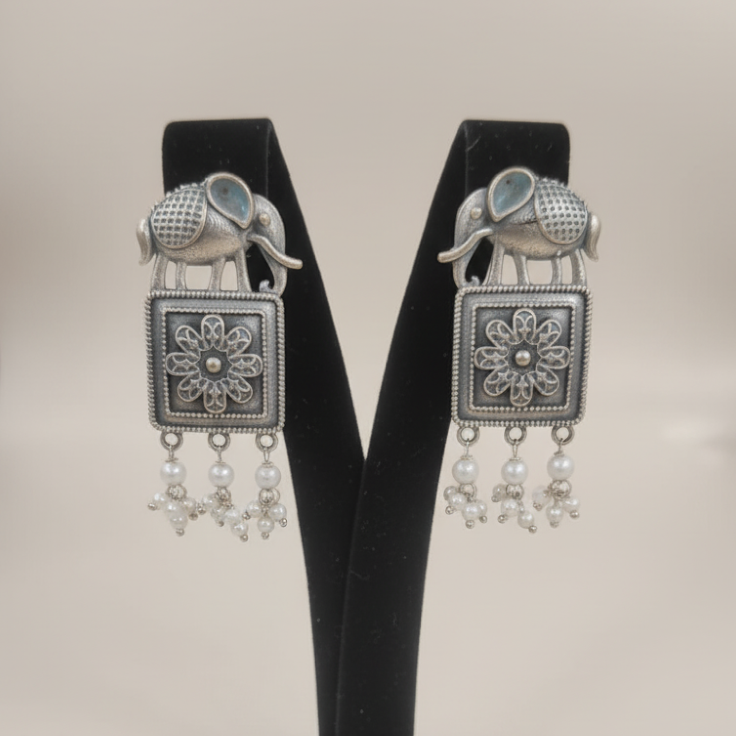 Oxidized silver earrings with elephant motifs, floral square design, and pearl drop detailing displayed on a black stand