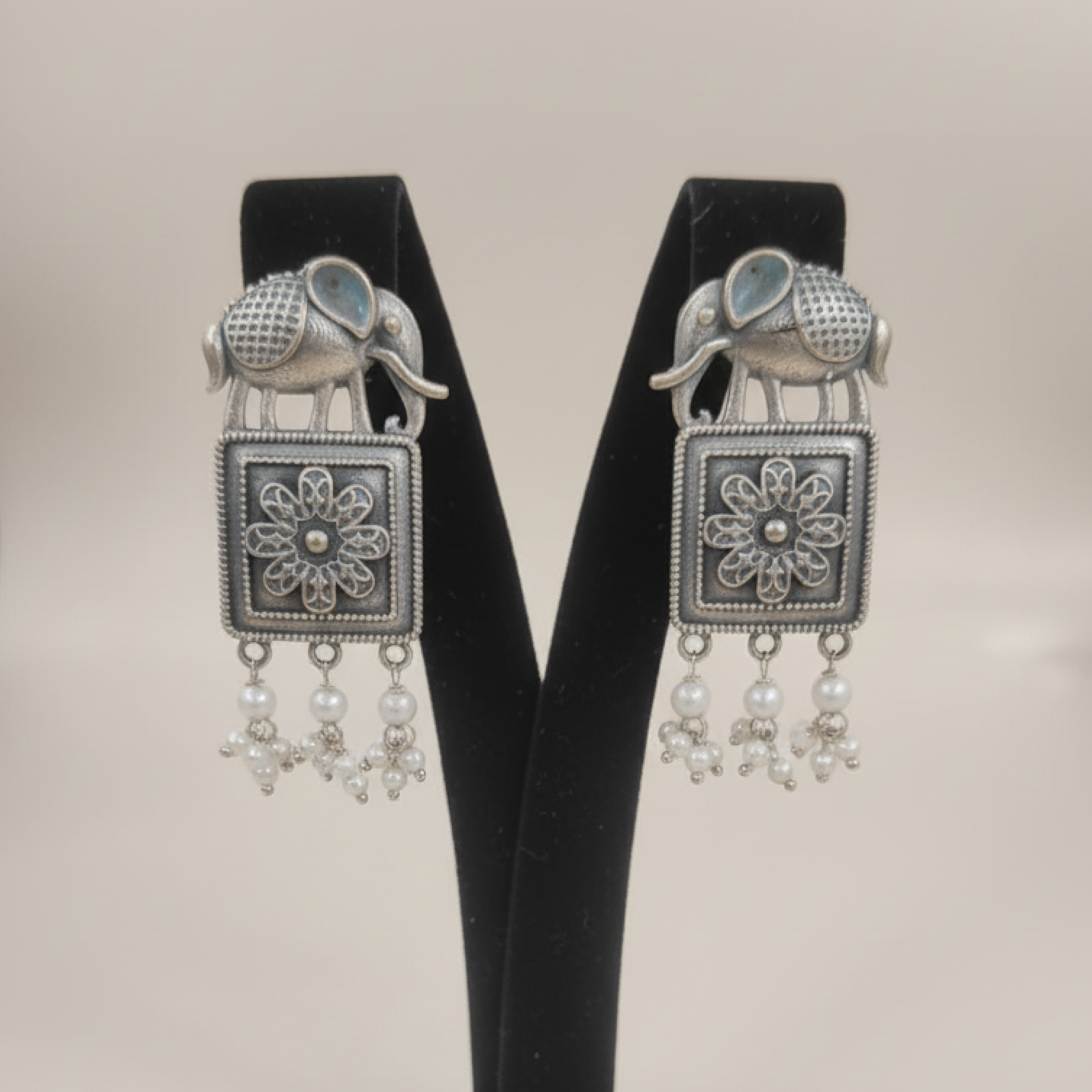 Oxidized silver earrings with elephant motifs, floral square design, and pearl drop detailing displayed on a black stand