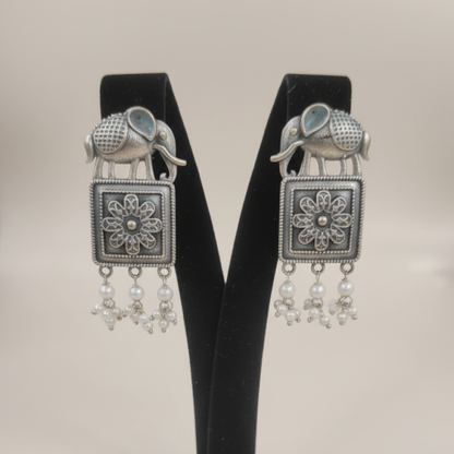 Oxidized silver earrings with elephant motifs, floral square design, and pearl drop detailing displayed on a black stand