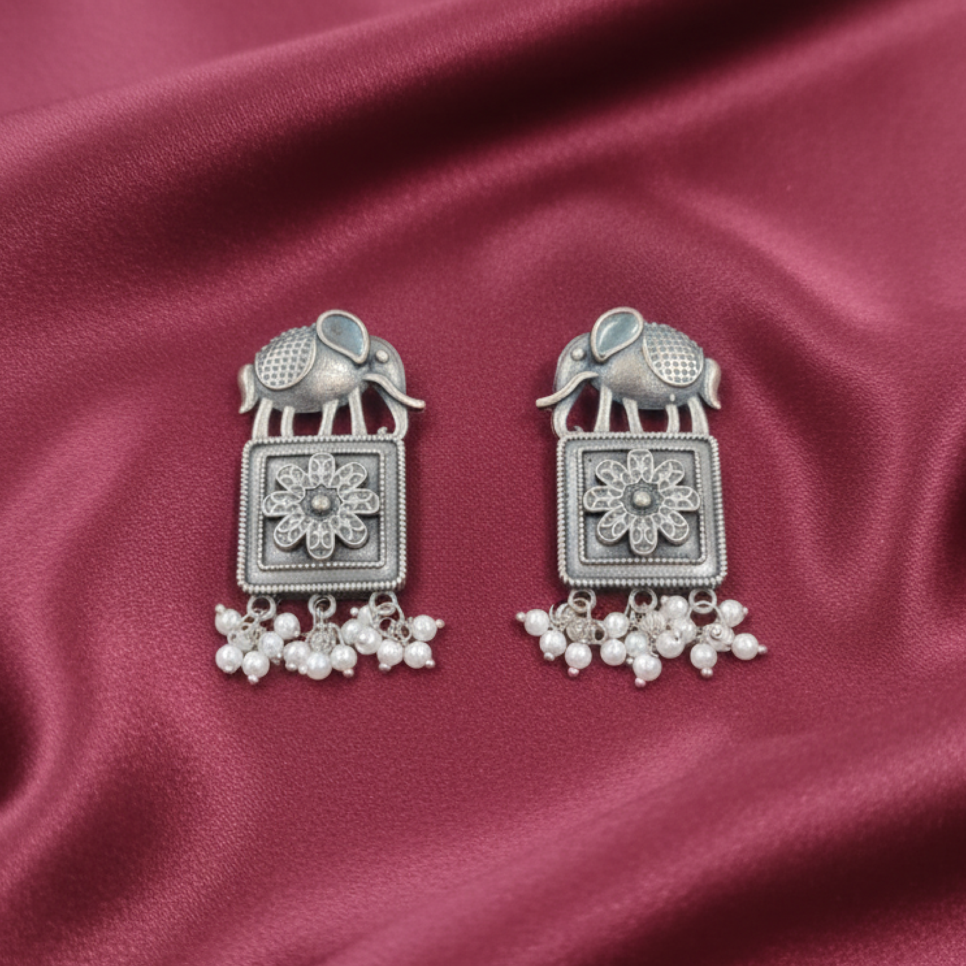 Oxidized silver earrings with elephant motifs, floral square design, and pearl drop detailing displayed on a black stand white