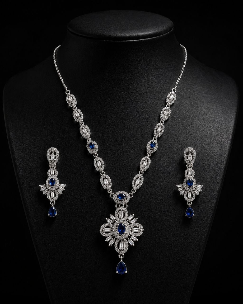 Elegant silver-plated necklace with large blue and clear stone pendant displayed on beige mannequin bust alongside matching blue teardrop earrings against soft beige fabric background 3