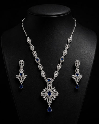 Elegant silver-plated necklace with large blue and clear stone pendant displayed on beige mannequin bust alongside matching blue teardrop earrings against soft beige fabric background 3