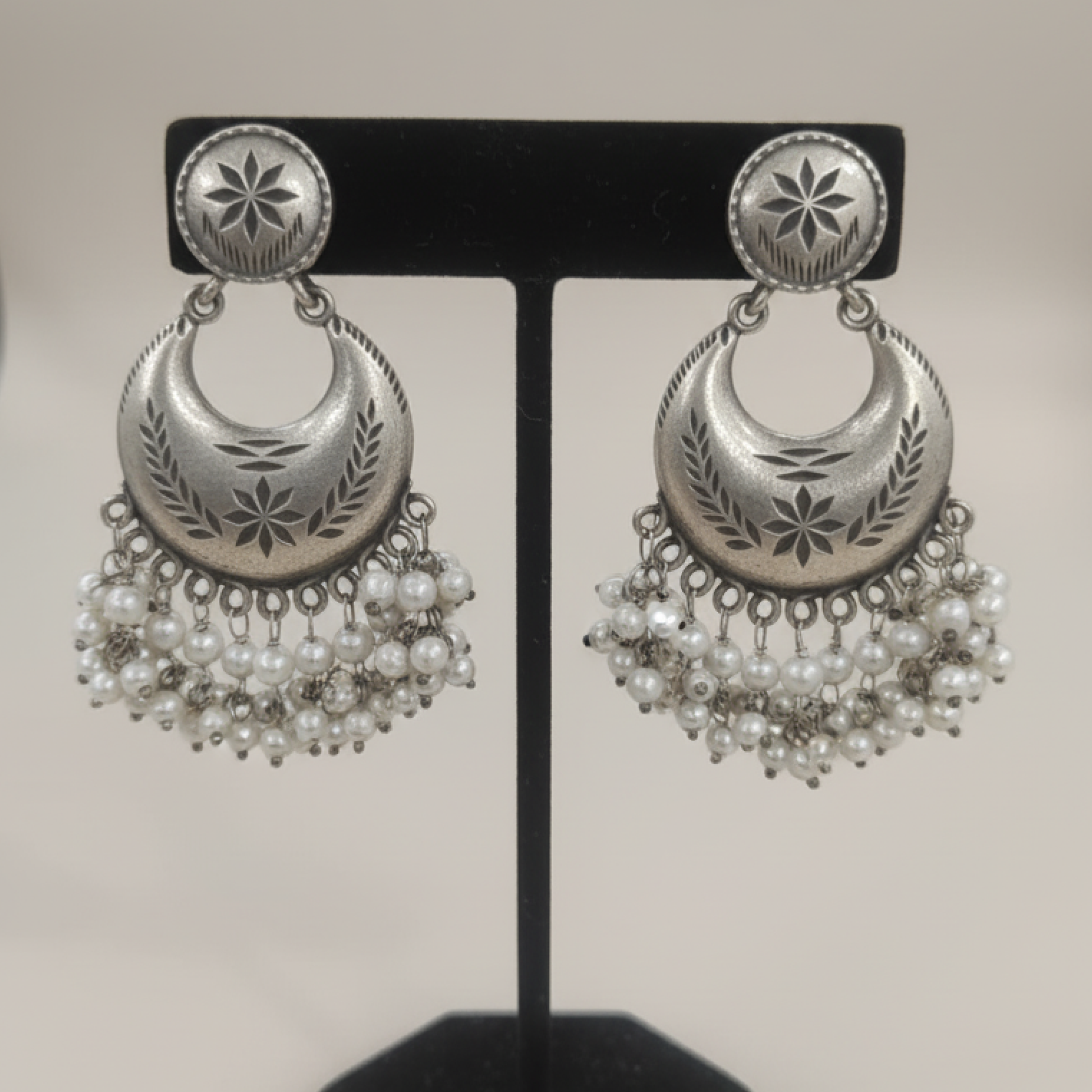 Oxidized silver crescent earrings with engraved detailing and pearl hangings displayed on a black stand